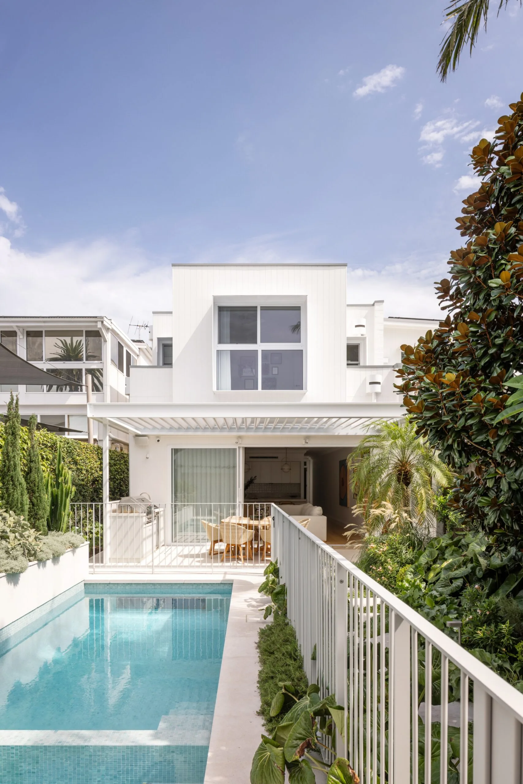 Modern white house with a balcony, pool, and lush garden under a blue sky.