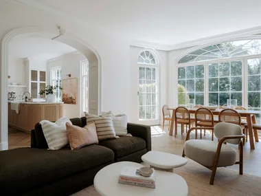 A bright living room with a sofa, armchair, coffee table, and dining area set by large windows.
