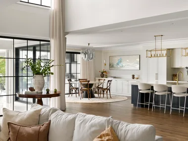 Modern open-plan living room and kitchen with large windows, light decor, and a dining area.