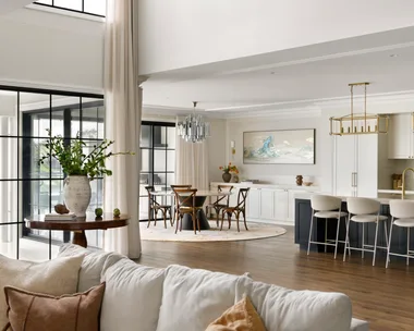 Modern open-plan living room and kitchen with large windows, light decor, and a dining area.