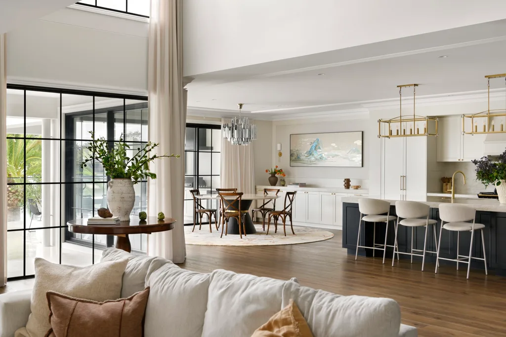Modern open-plan living room and kitchen with large windows, light decor, and a dining area.