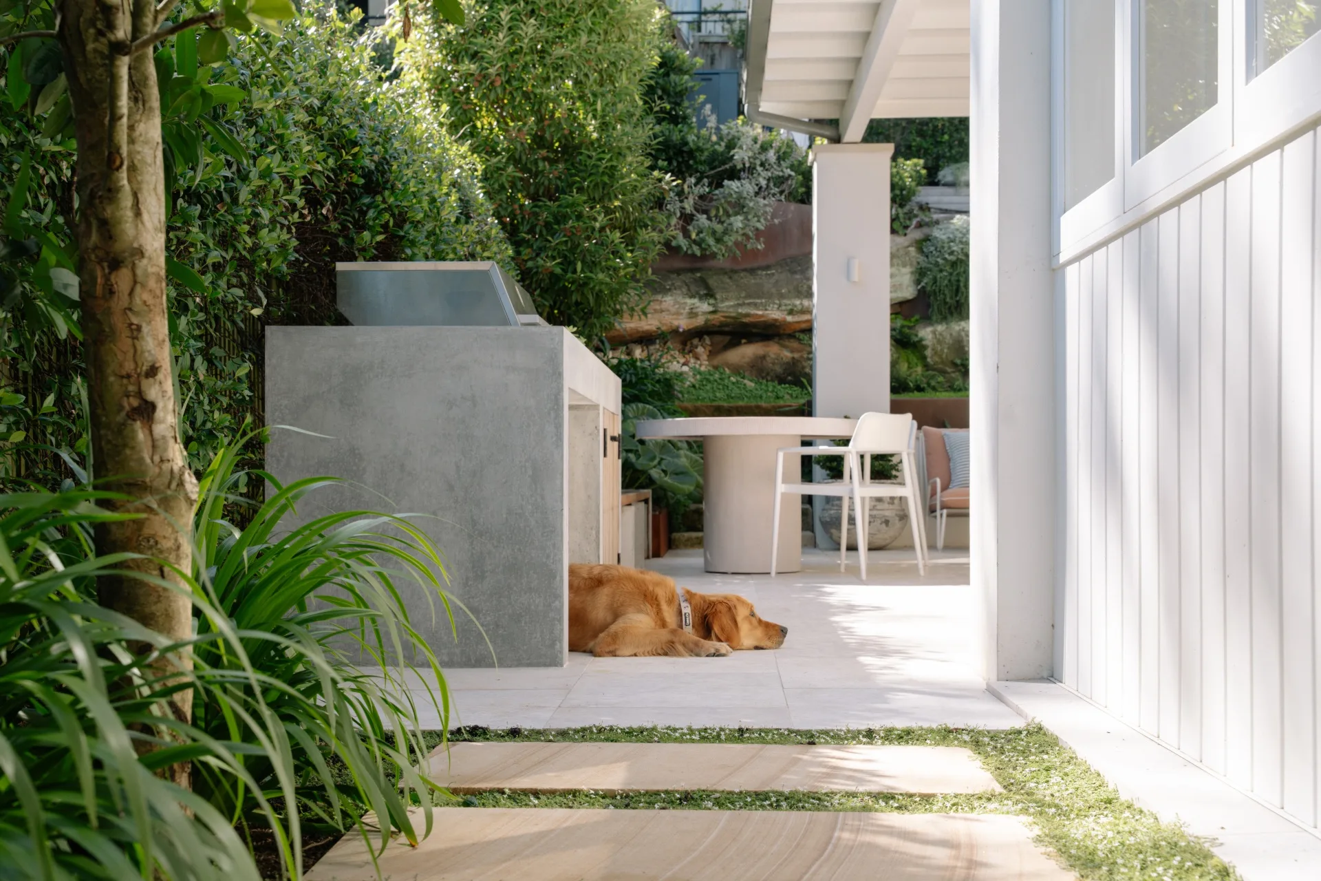 Golden retriever lying on a patio beside an outdoor kitchen surrounded by lush greenery and a table with chairs.