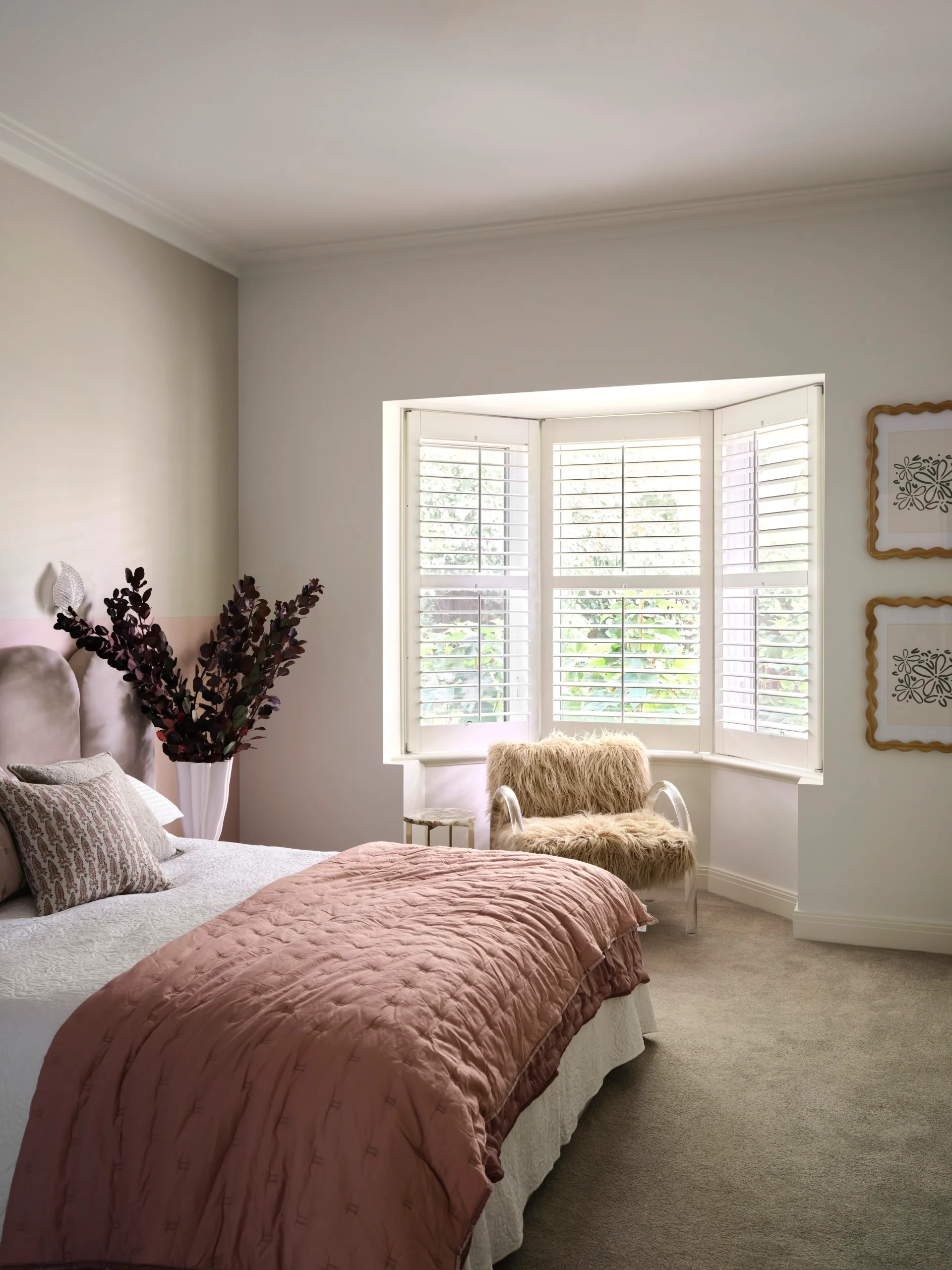 Bedroom with a pink bedspread, bay window with white shutters, and a faux fur chair in the corner.