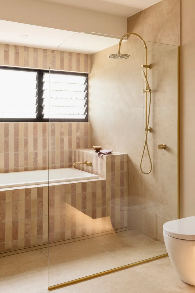 Luxurious bathroom with glass shower, gold fixtures, striped tiles, bathtub under a louvered window, and beige walls.