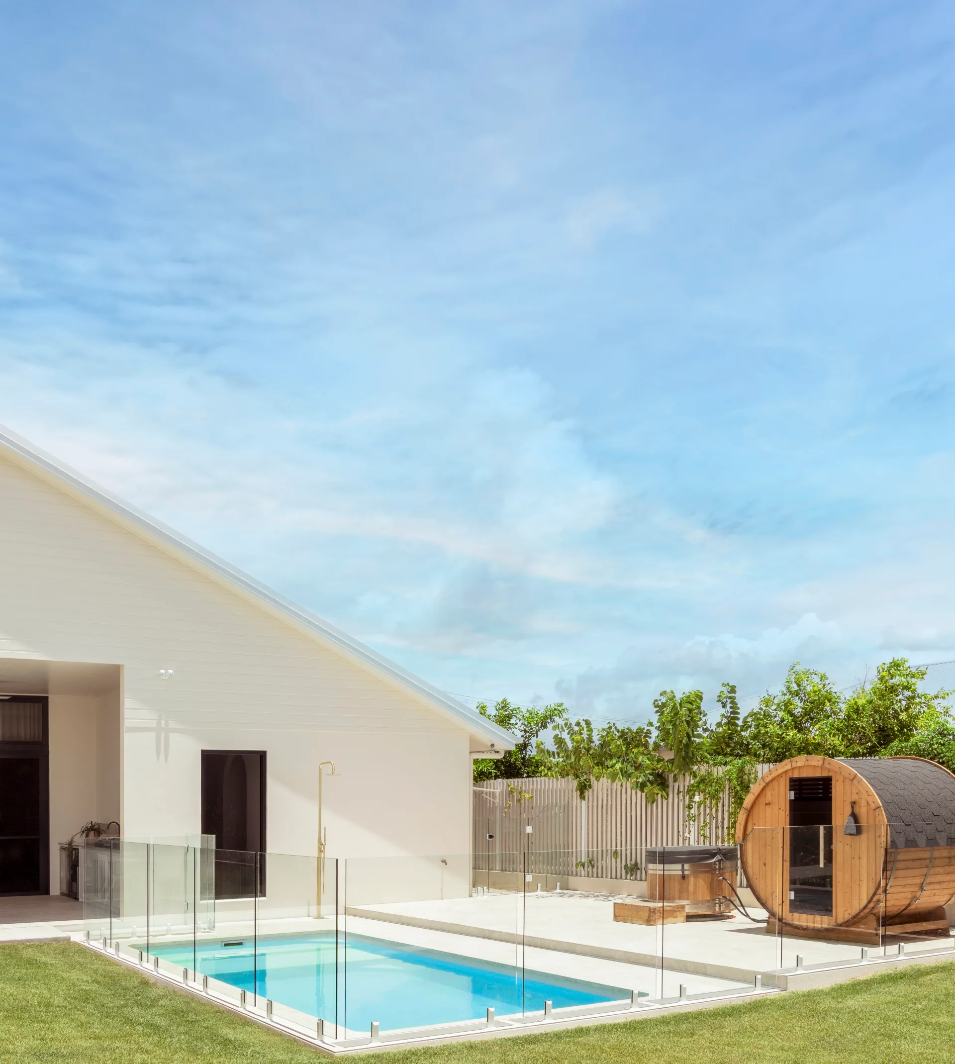 Outdoor modern patio with glass-fenced pool, a wooden sauna, and a white house under a clear blue sky.