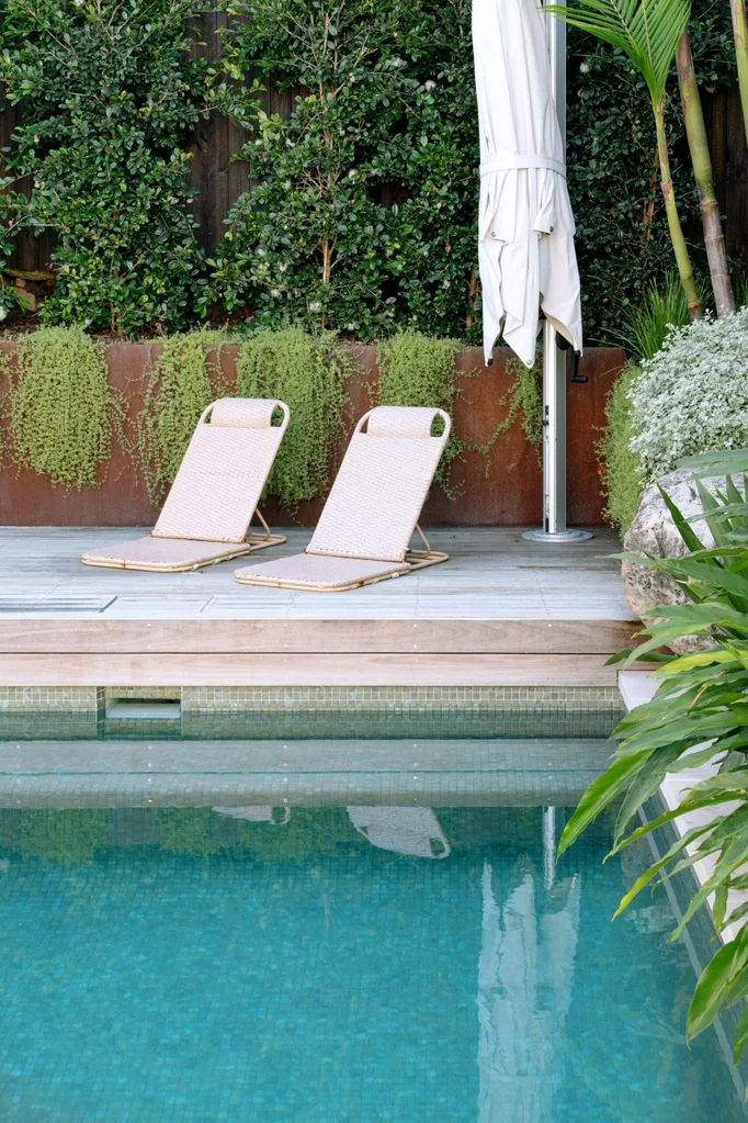 Two lounge chairs and a closed umbrella by a pool, surrounded by lush greenery and a wooden deck.