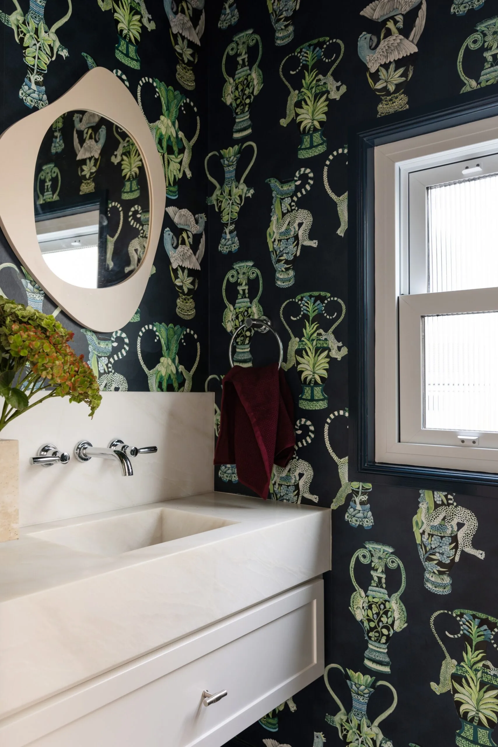 Dark-themed bathroom with leafy and animal print wallpaper, a modern white sink, round mirror, and a small window.