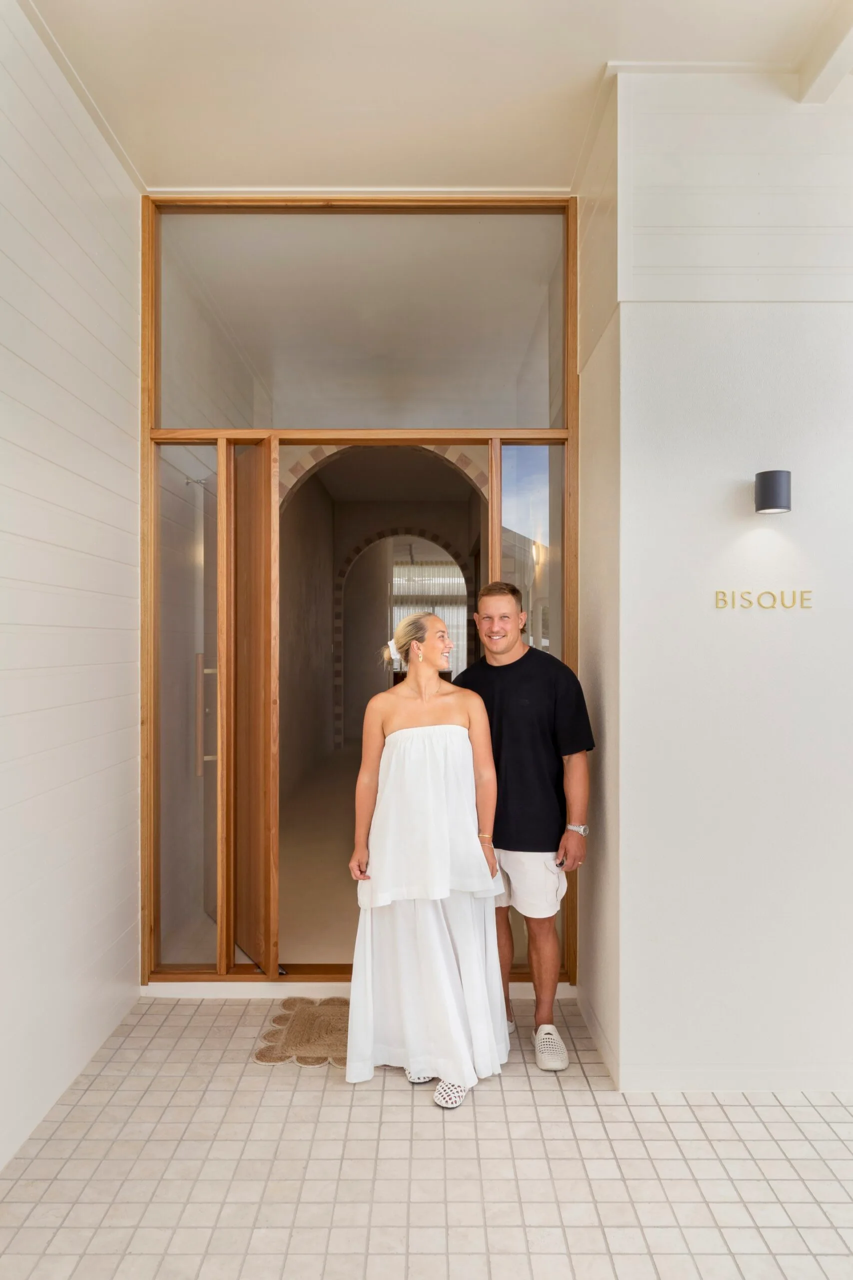 Couple smiling at each other outside a building labeled "BISQUE," with a wooden entrance and tiled floor.