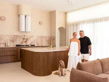 Couple in a modern kitchen with marble countertops, a dog sits by their feet, and a brown sofa is in the foreground.