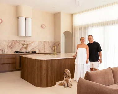 Couple in a modern kitchen with marble countertops, a dog sits by their feet, and a brown sofa is in the foreground.