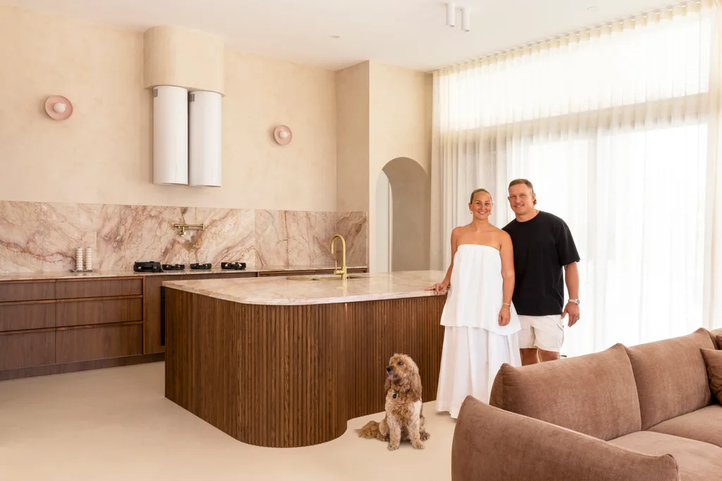 Couple in a modern kitchen with marble countertops, a dog sits by their feet, and a brown sofa is in the foreground.