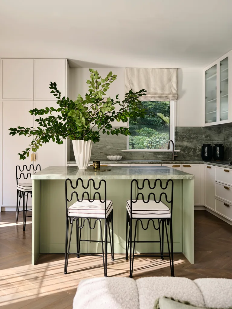 Green kitchen island with decorative chairs, a large leafy plant in a vase, and modern white cabinetry with a window view.