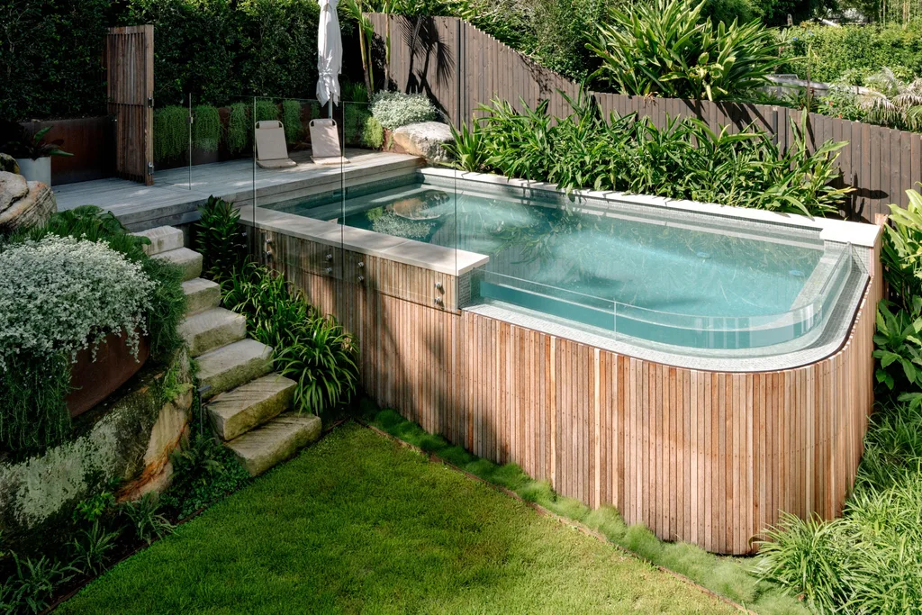 Elevated backyard pool with wooden paneling, glass fence, and lush surrounding greenery.