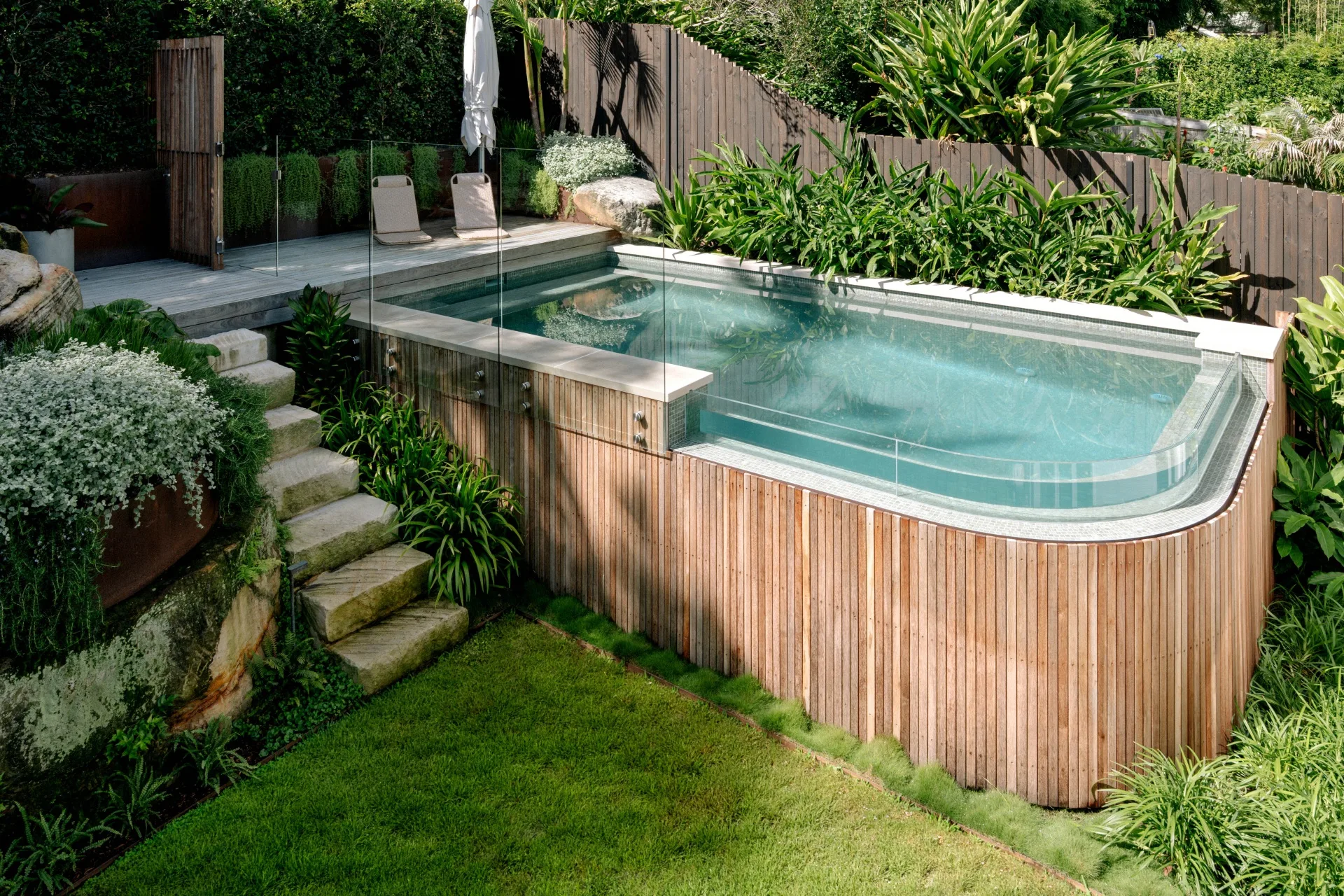 Elevated backyard pool with wooden paneling, glass fence, and lush surrounding greenery.