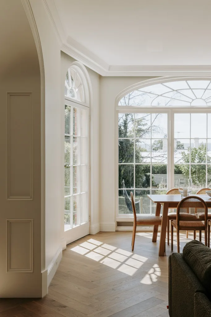 Bright room with large arched windows, wooden dining table, chairs, and sunlight streaming onto the wooden floor.