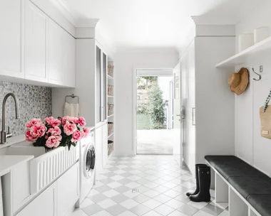 A white laundry and mudroom with chequered grey and white floor tiles and wall mosaics, a white butler's sink, bench seat, drying cupboard and storage.