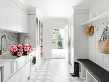 A white laundry and mudroom with chequered grey and white floor tiles and wall mosaics, a white butler's sink, bench seat, drying cupboard and storage.