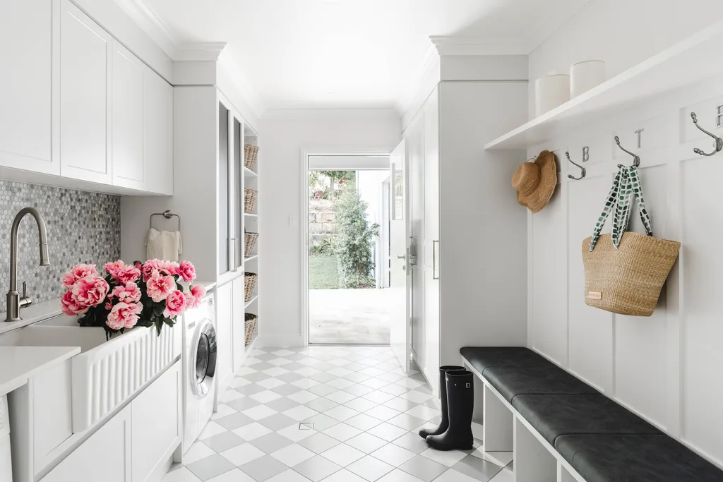 A white laundry and mudroom with chequered grey and white floor tiles and wall mosaics, a white butler's sink, bench seat, drying cupboard and storage.