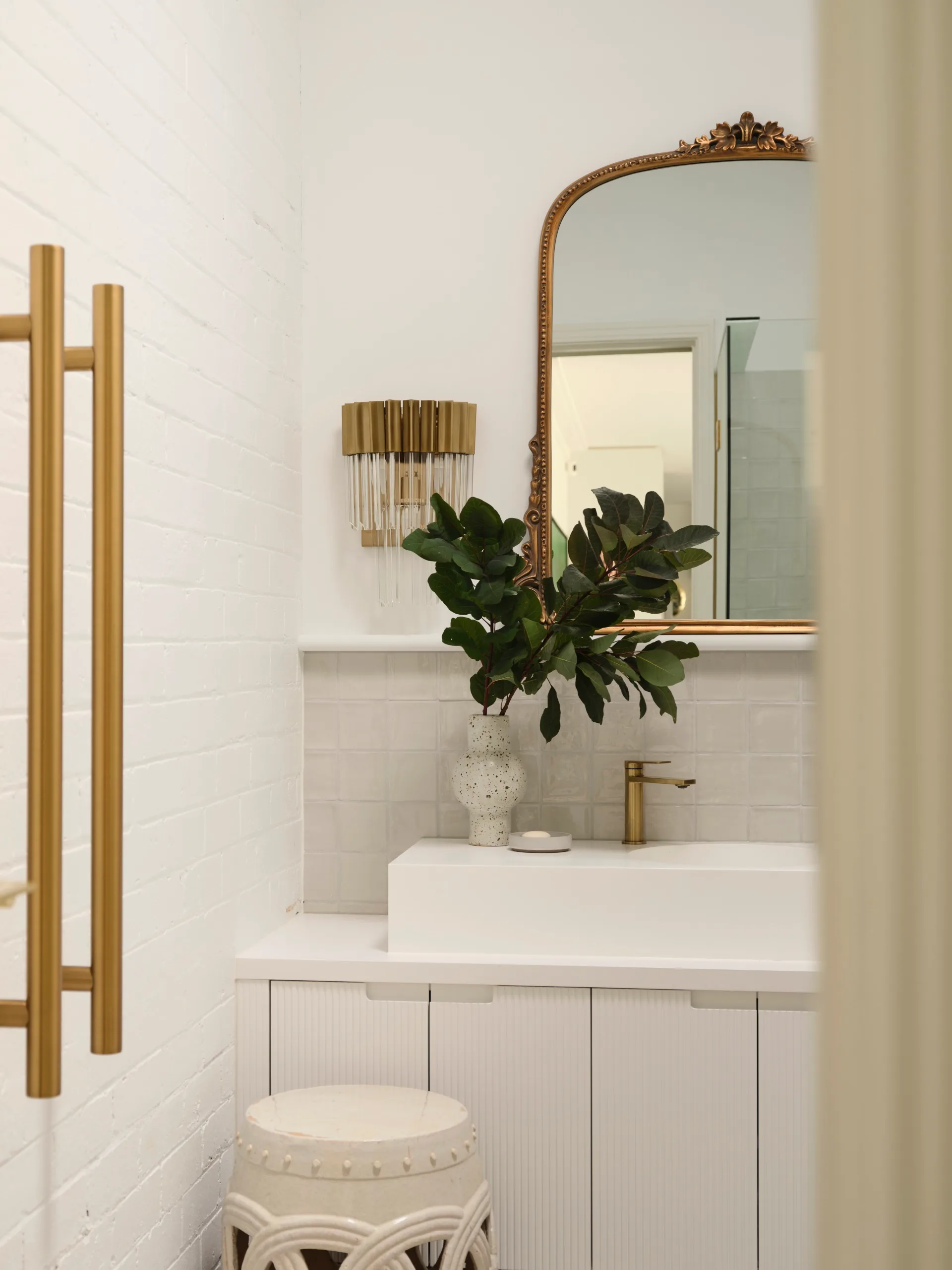 Elegant bathroom with white sink, gold faucet, ornate mirror, and a vase with leafy greens on a white vanity.