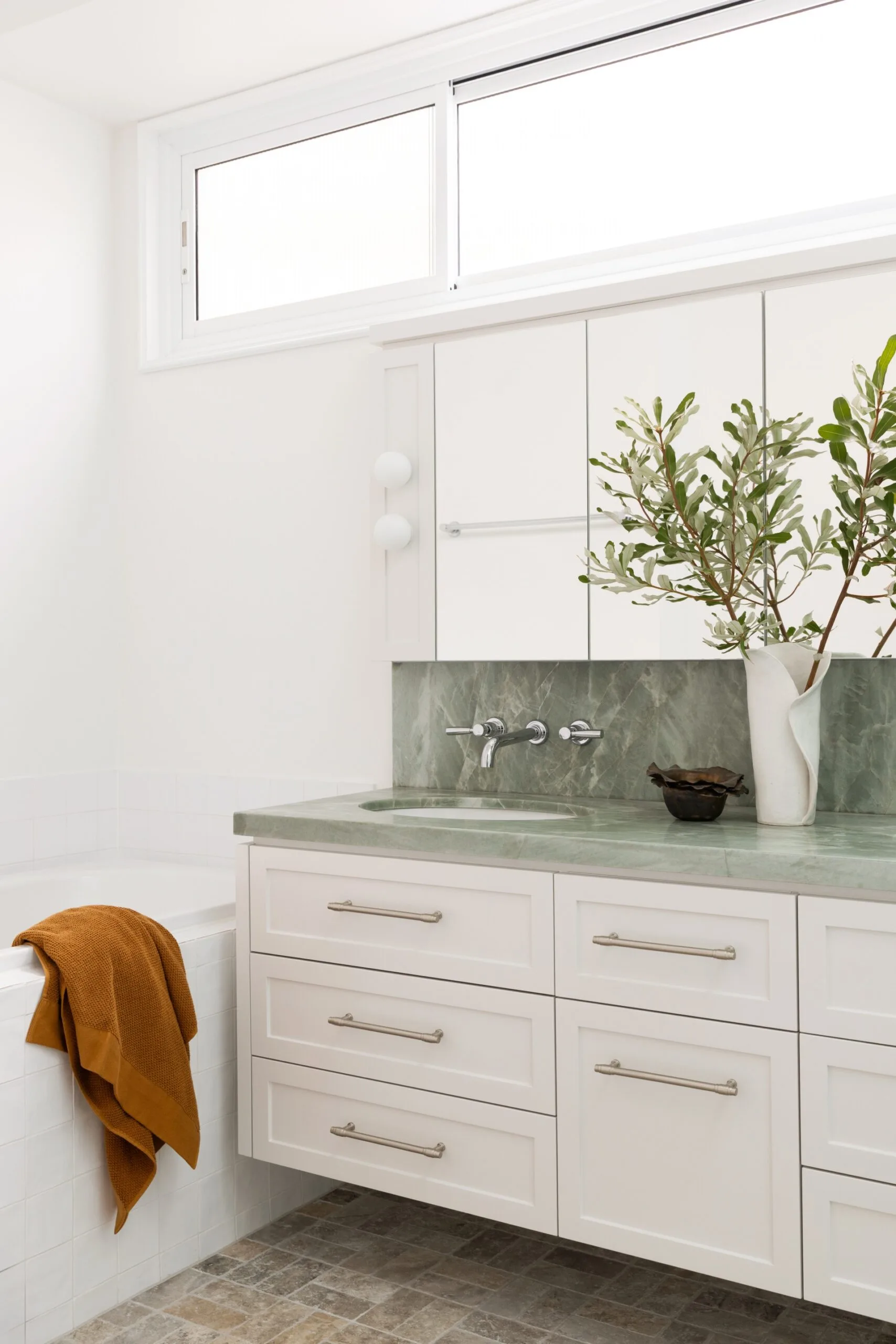 White bathroom with green marble countertop, round mirror, and a white vase with green branches. Orange towel on tub.
