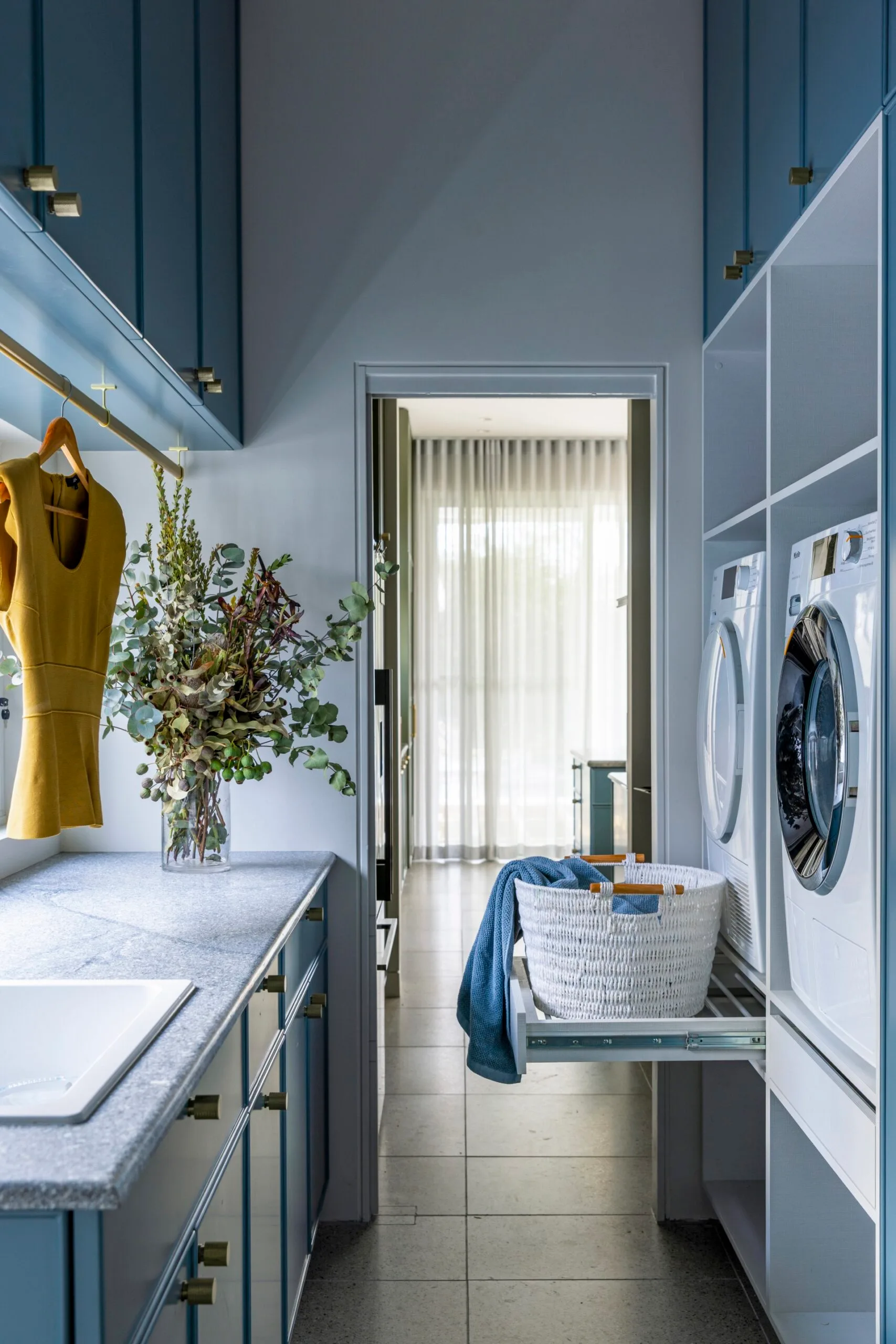 A sophisticated blue laundry with a narrow galley layout fitted with overhead and under-bench cupboards, open shelving and pull-out drying racks below the washing machine and dryer, plus a yellow shirt hanging from a brass drying rail.