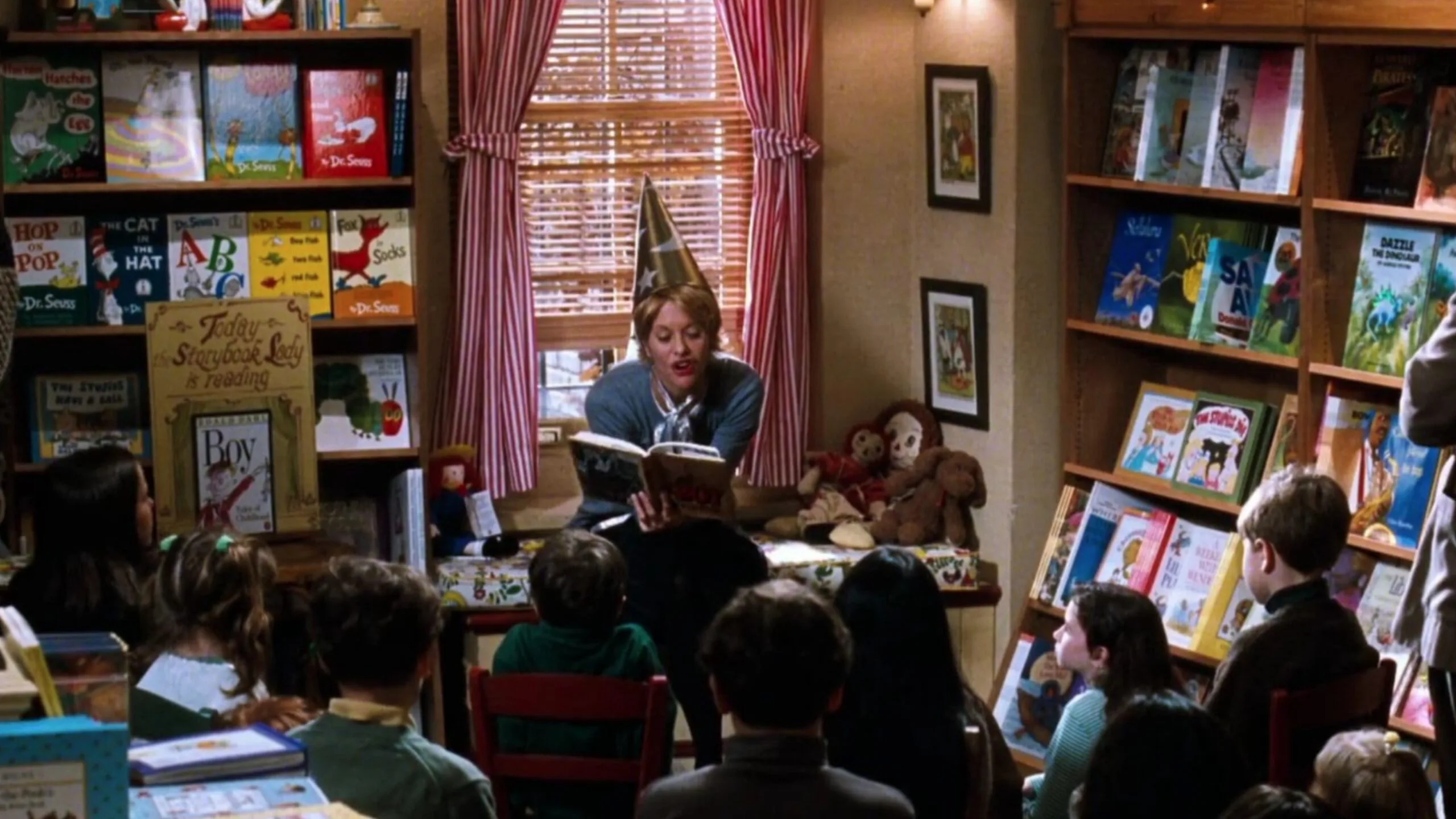 Still from the movie 'You've Got Mail' depicting Meg Ryan's character, Kathleen Kelly. Storytime at a bookstore with Meg Ryan in a wizard hat reading to children, surrounded by colorful books and toys. Movie directed by Nora Ephron.