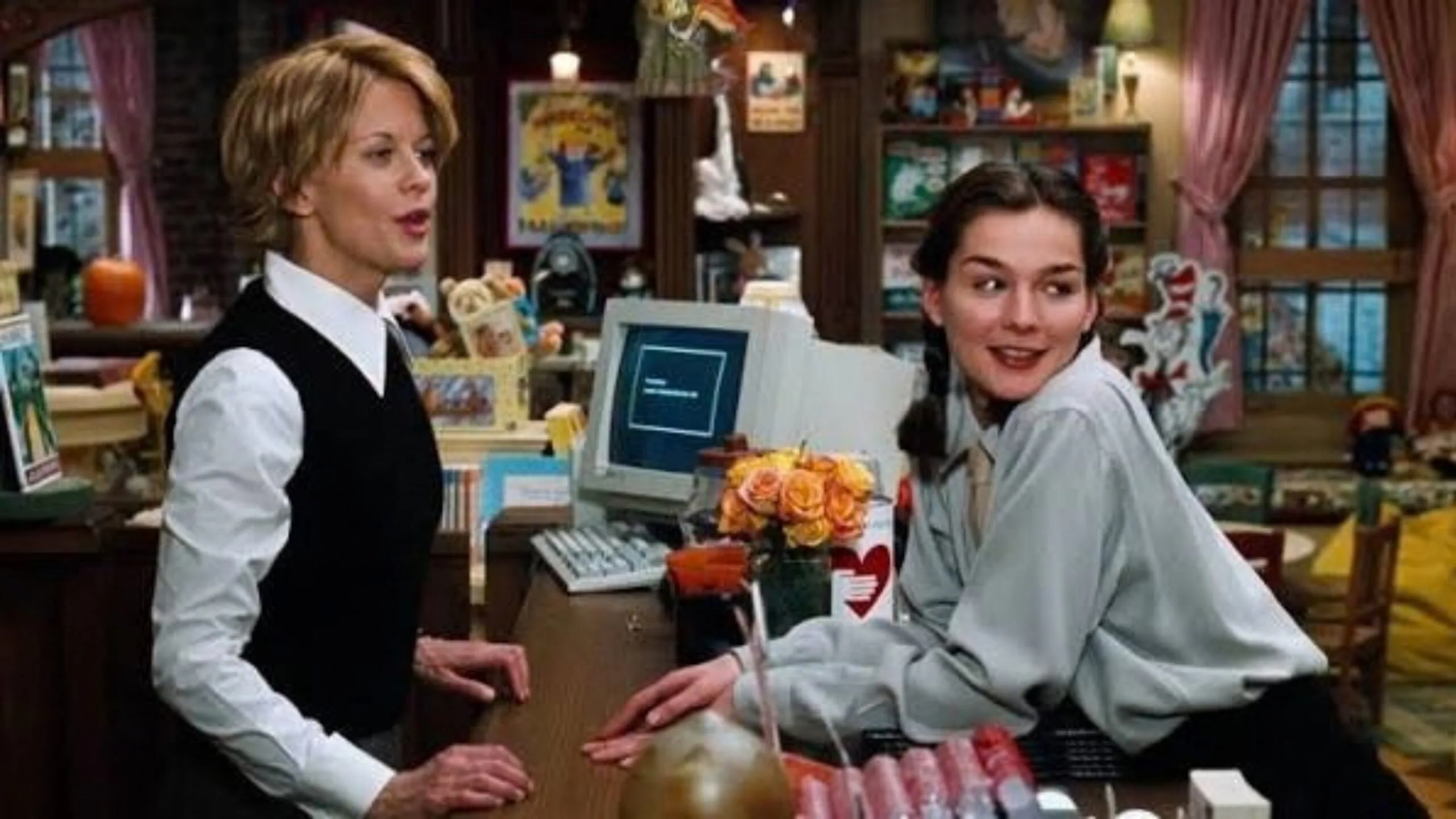 Still from the movie 'You've Got Mail' depicting Meg Ryan's character, Kathleen Kelly. She is standing with another woman in a cosy bookstore, one standing and one leaning on a counter with a boxy late-90s computer. Movie directed by Nora Ephron.