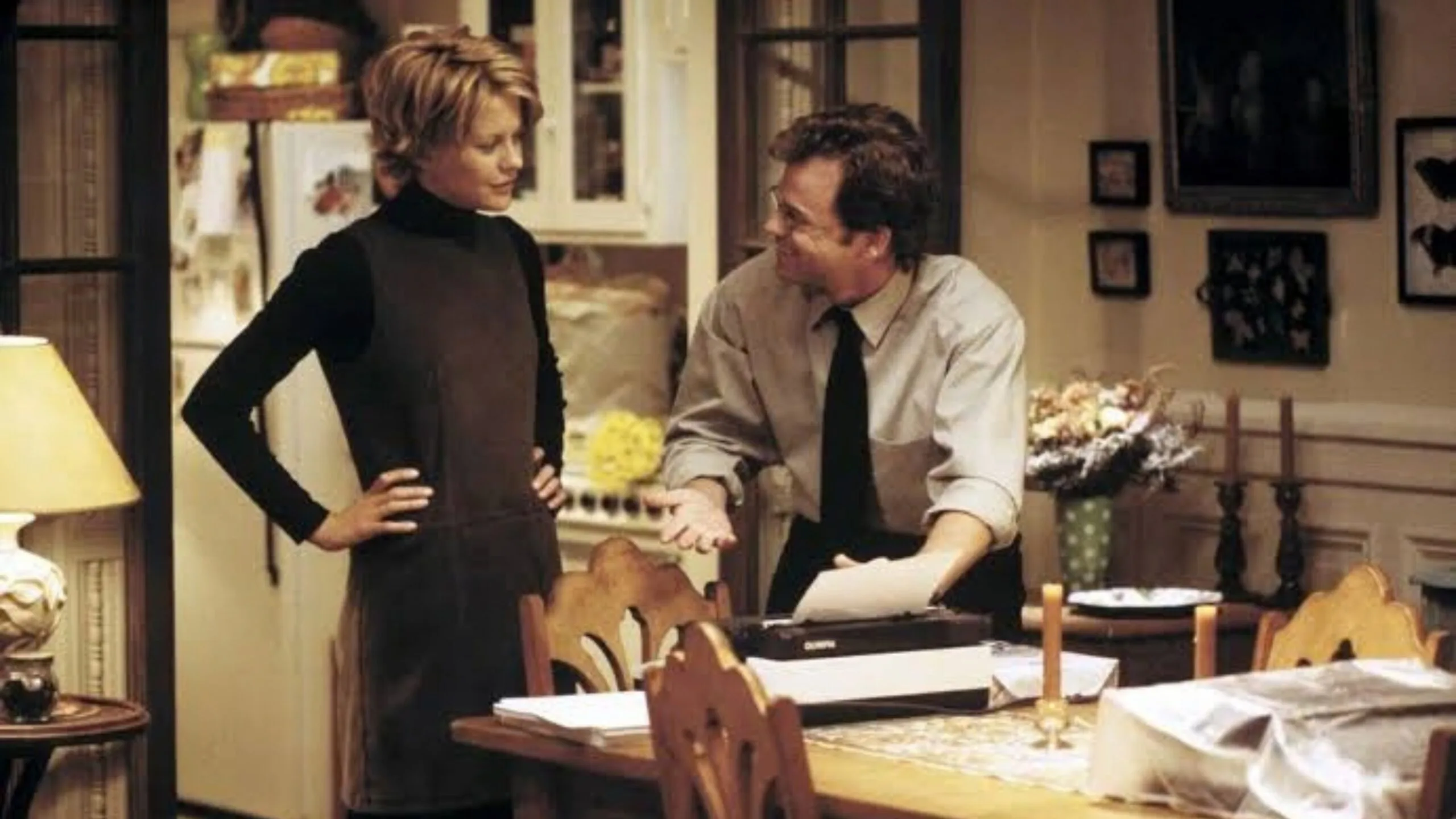 Still from the movie 'You've Got Mail' depicting the apartment of Meg Ryan's character, Kathleen Kelly. She and a man are talking in a cosy kitchen and dining room, standing near a dining table with papers and a typewriter. Movie directed by Nora Ephron.