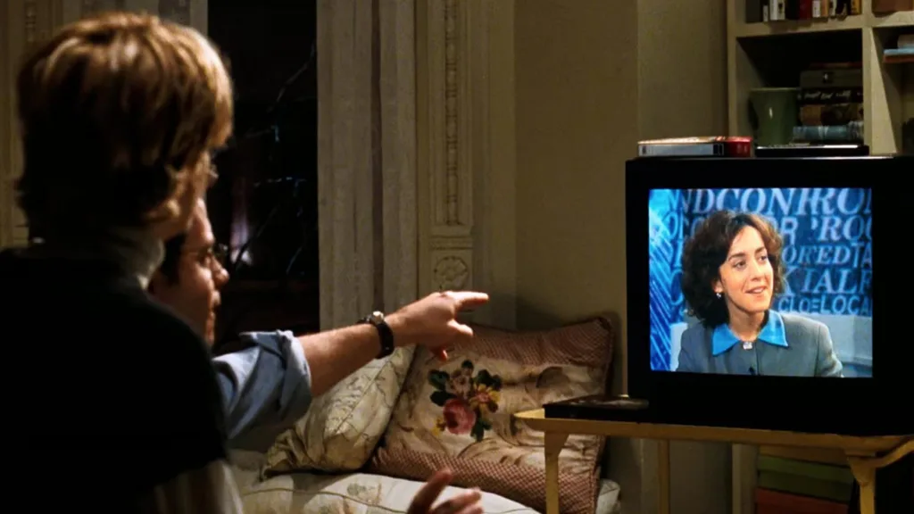 Still from the movie 'You've Got Mail' depicting the apartment of Meg Ryan's character, Kathleen Kelly. A man sitting beside her on a couch points at a TV screen showing a woman speaking. A floral cushion is on the sofa nearby and a cluttered bookshelf is in the background. Movie directed by Nora Ephron.