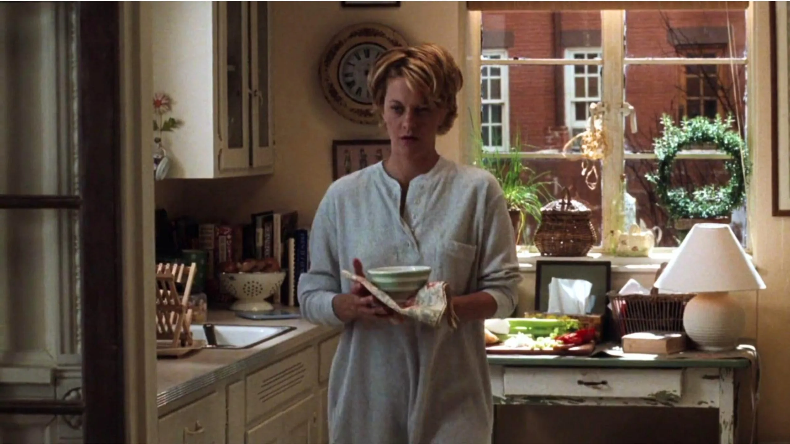 Still from the movie 'You've Got Mail' depicting the apartment of Meg Ryan's character, Kathleen Kelly. She stands in a shabby chic and cosy kitchen holding a bowl, with shelves and a window in the background. Movie directed by Nora Ephron.