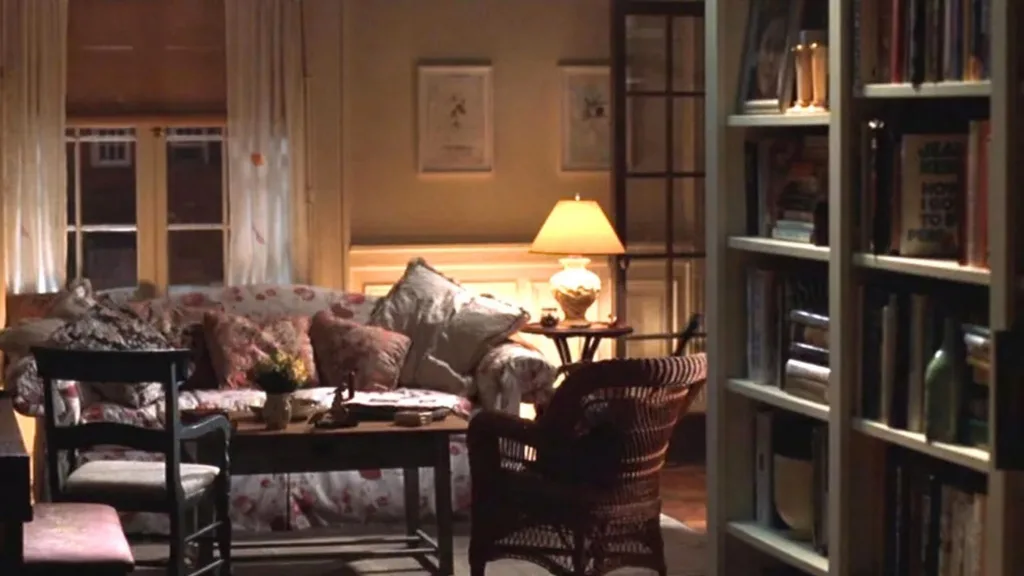 Still from the movie 'You've Got Mail' depicting the apartment of Meg Ryan's character, Kathleen Kelly. Cozy living room with floral sofa, cushions, a wicker chair, and a lit lamp on a side table, next to a bookshelf. Movie directed by Nora Ephron.