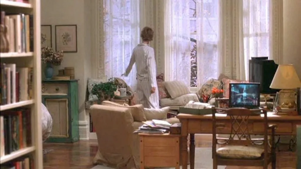 Still from the movie 'You've Got Mail' depicting the apartment of Meg Ryan's character, Kathleen Kelly. Interior of a cosy living room with Meg Ryan in pajamas looking out a large window; a cluttered desk and full bookshelf visible. Movie directed by Nora Ephron.