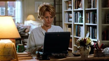 Still from the movie 'You've Got Mail' depicting the apartment of Meg Ryan's character, Kathleen Kelly. She is in a cosy room using a laptop at a wooden desk, surrounded by books, flowers, and a lamp. Movie directed by Nora Ephron.