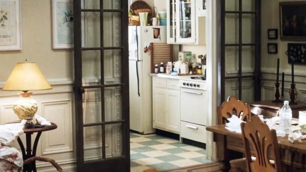 Still from the movie 'You've Got Mail' depicting the apartment of Meg Ryan's character, Kathleen Kelly. Cosy kitchen with white cabinets, checkered floor, dining table, and glass-paned doors, softly lit by a table lamp. Movie directed by Nora Ephron.