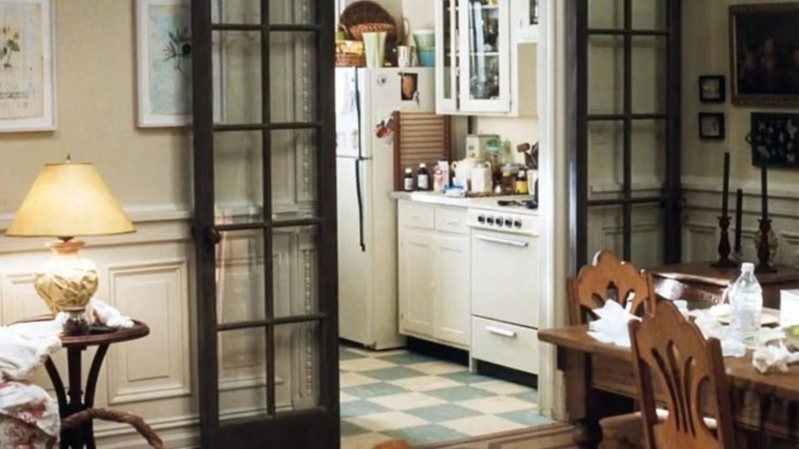 Still from the movie 'You've Got Mail' depicting the apartment of Meg Ryan's character, Kathleen Kelly. Cosy kitchen with white cabinets, checkered floor, dining table, and glass-paned doors, softly lit by a table lamp. Movie directed by Nora Ephron.