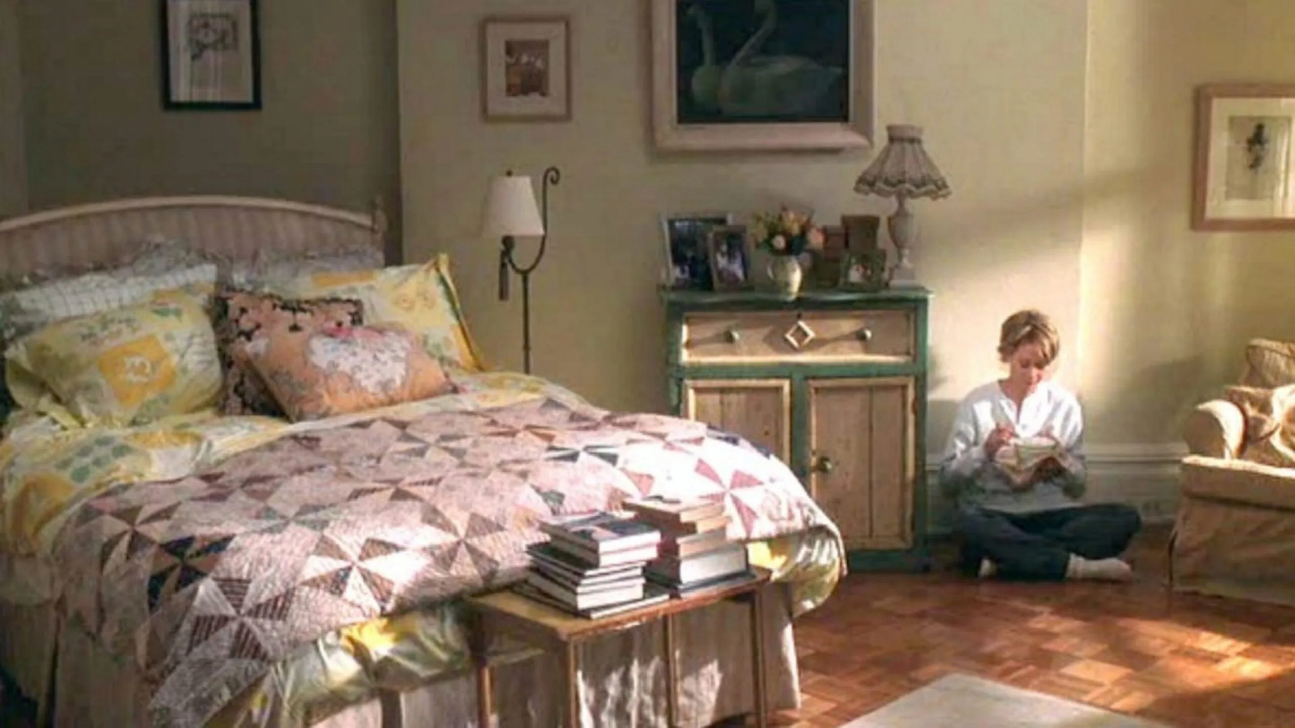 Still from the movie 'You've Got Mail' depicting the apartment of Meg Ryan's character, Kathleen Kelly. She is sitting cross legged on the timber floor beside her bed in a cozy bedroom with patterned quilt and stacked books. Movie directed by Nora Ephron.