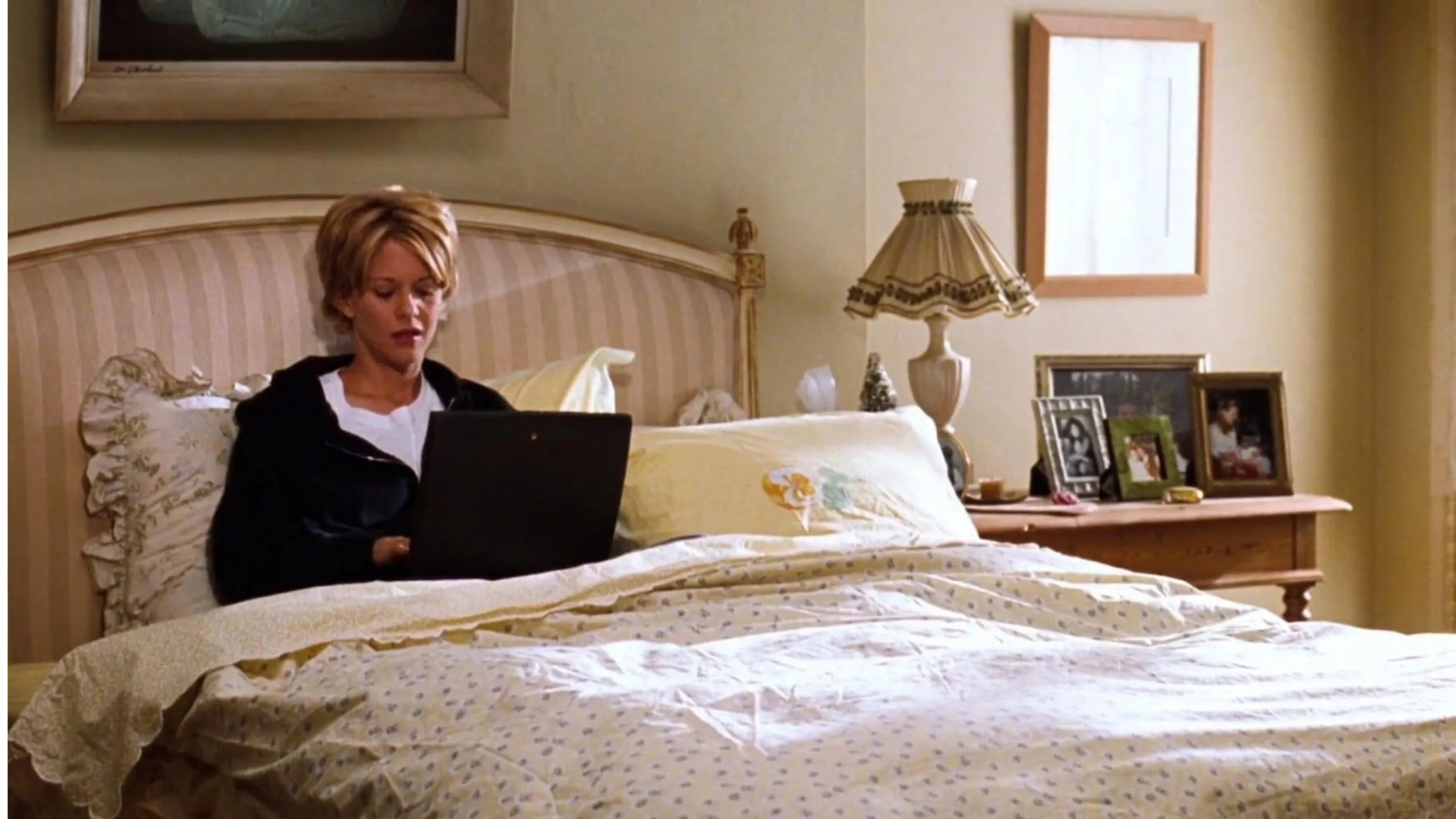 Still from the movie 'You've Got Mail' depicting the apartment of Meg Ryan's character, Kathleen Kelly. She is sitting in bed using a laptop; bedroom with bedside table, framed photos, and a lamp. Movie directed by Nora Ephron.