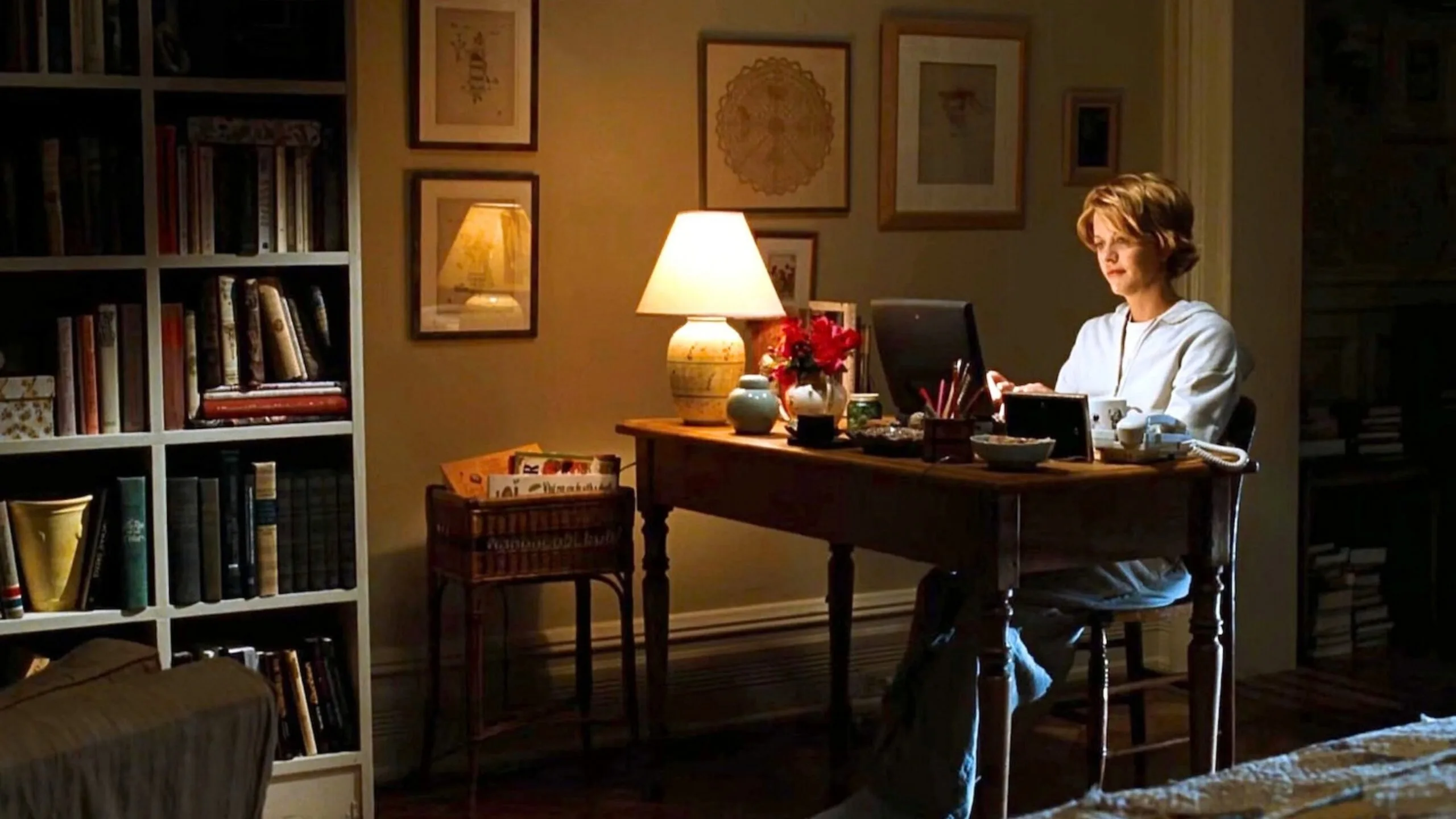 Still from the movie 'You've Got Mail' depicting the apartment of Meg Ryan's character, Kathleen Kelly. She is in her living room and study, typing on a laptop at a wooden desk, surrounded by books and a lit table lamp. There is framed art on the wall beside her. Movie directed by Nora Ephron.