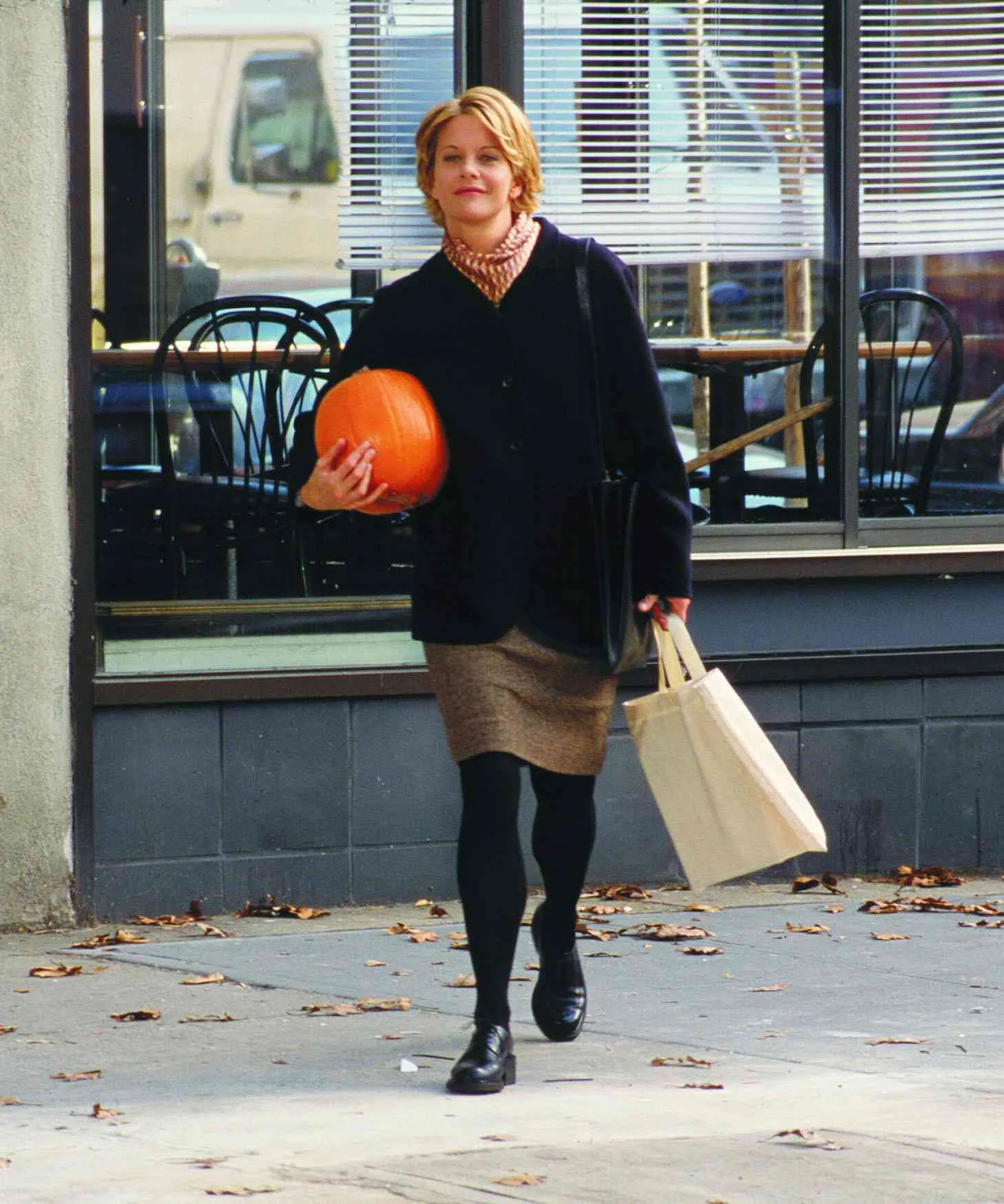 Still from the movie 'You've Got Mail' depicting Meg Ryan in autumn clothing walking on leaf-strewn sidewalk carrying a pumpkin and a tote bag, with a cafe in the background. Movie directed by Nora Ephron.