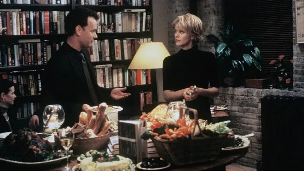 Still from the movie 'You've Got Mail' depicting Meg Ryan's character, Kathleen Kelly, and Tom Hanks as Joe Fox. They stand in an apartment and converse over a table filled with food, surrounded by a cozy, lamp-lit atmosphere. A bookshelf filled with books is in the background, beside a brick wall. Movie directed by Nora Ephron.