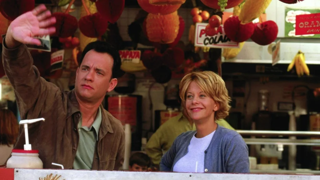 Still from the movie 'You've Got Mail' depicting Meg Ryan's character, Kathleen Kelly and Tom Hanks as Joe Fox. They are standing at a market stall, one waving, with paper decorations in the shape of fruit hanging above them. Movie directed by Nora Ephron.
