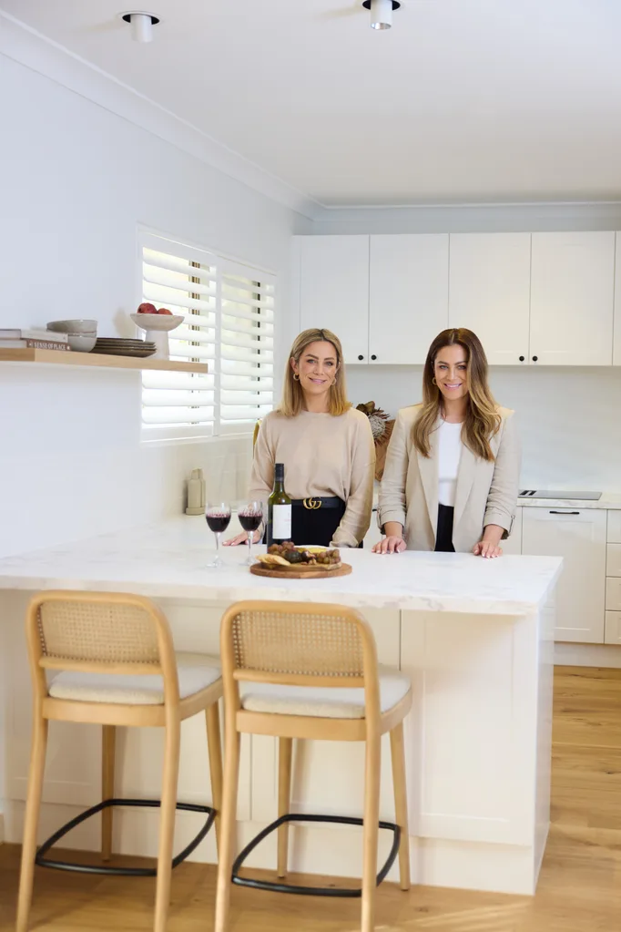 Alisa and Lysandra Fraser stand at a kitchen island with wine and snacks, inside the modern white kitchen they renovated for their mum.