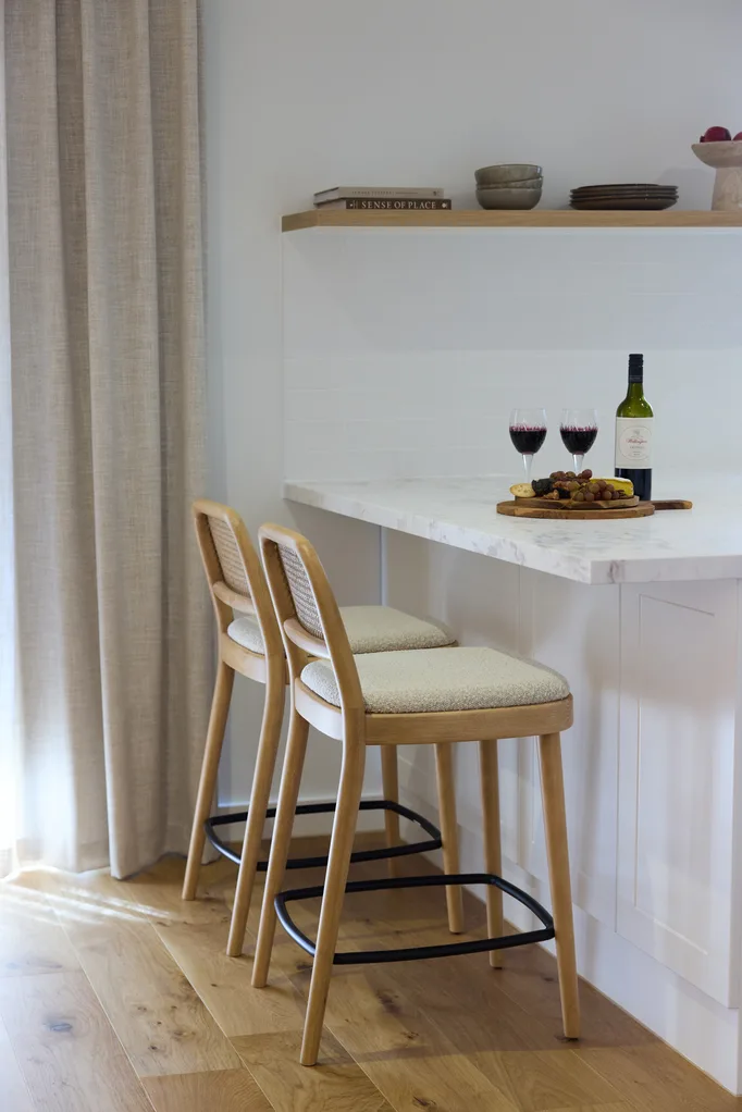 Two wooden bar stools at a marble countertop with wine bottle, two glasses, and a cheese platter.