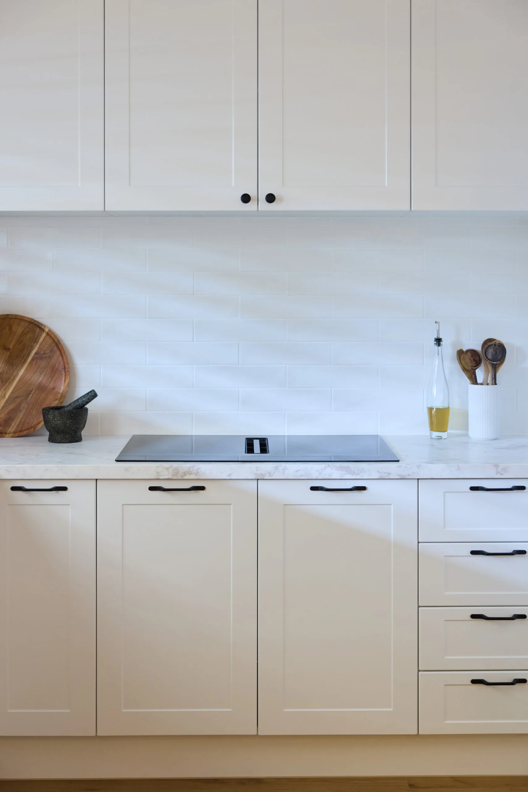 Sleek white kitchen cabinets with black handles, a built-in cooktop, and utensils on a marble countertop.