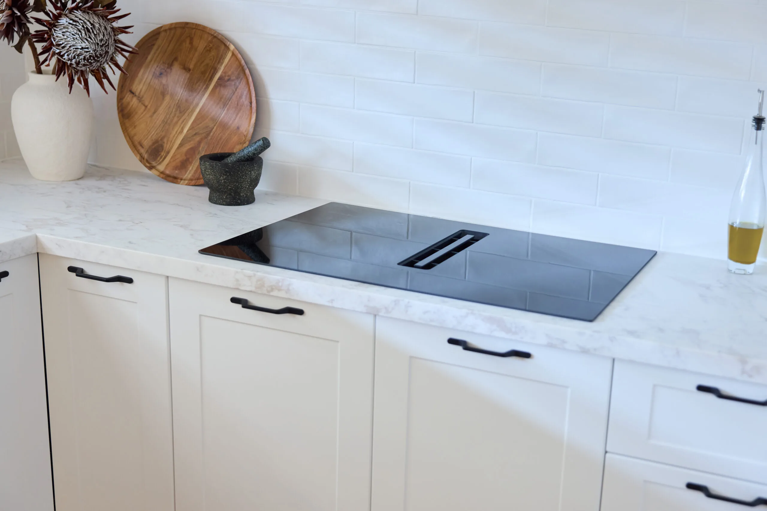 Modern kitchen countertop with vase, wooden board, mortar, and induction stove. White cabinets and backsplash.