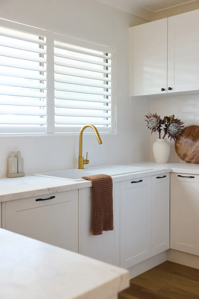 Modern kitchen with white cabinets, gold faucet, brown towel, and natural decor in vase on counter.