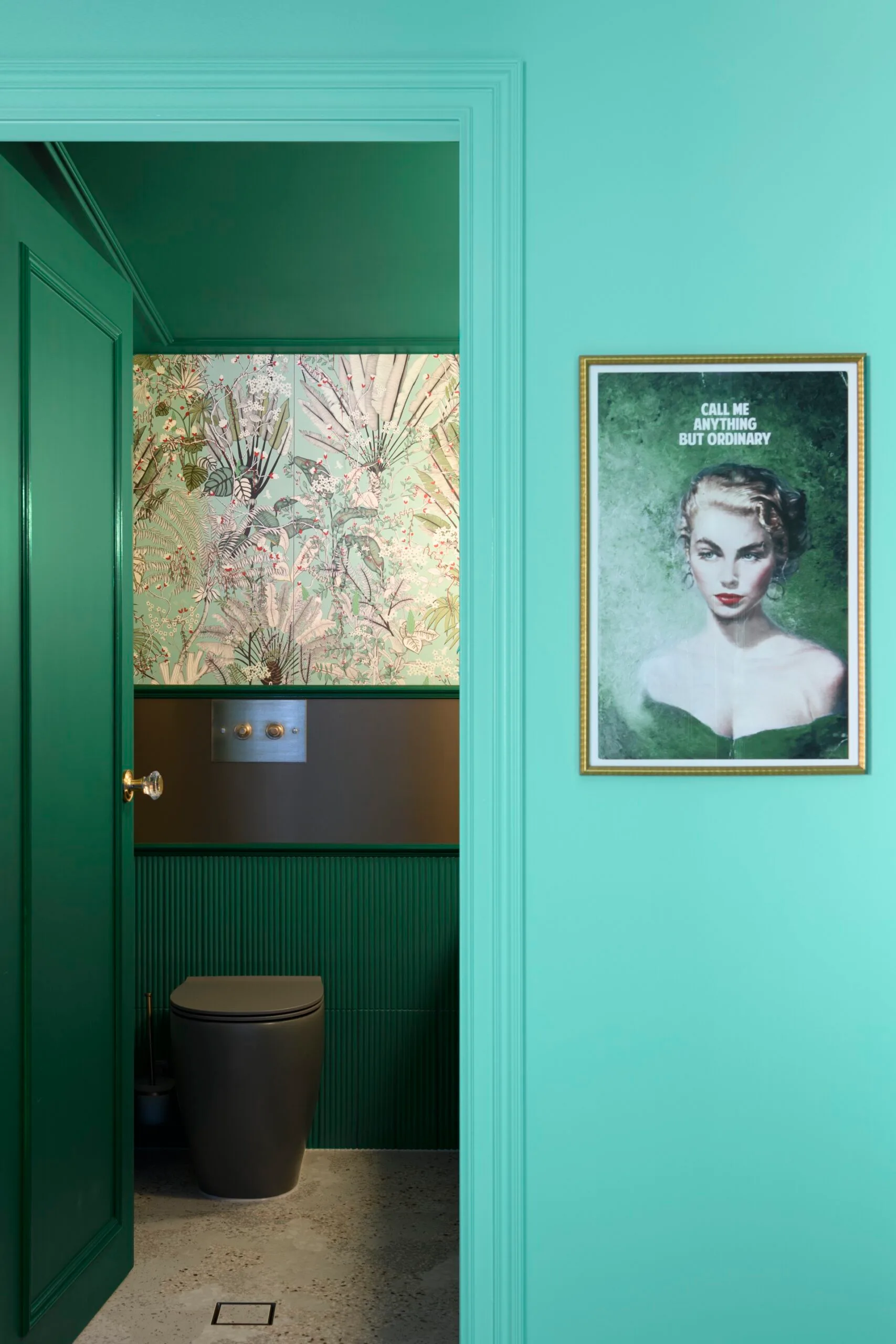 A teal bathroom with floral wallpaper, black toilet, and a framed portrait of a woman labeled "Call Me Anything But Ordinary."