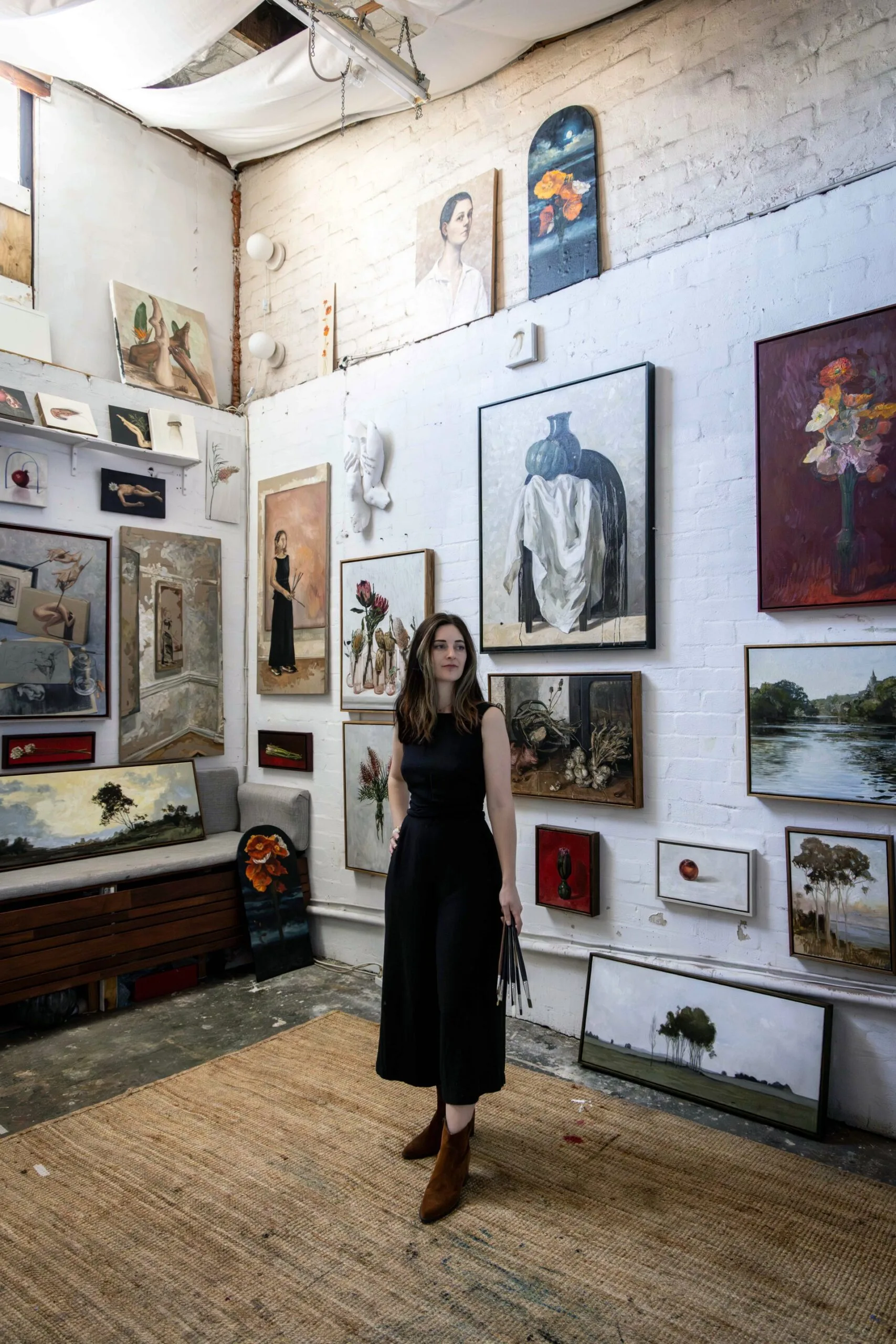 Artist in a studio surrounded by various paintings on white brick walls, holding paintbrushes, standing on a woven rug.