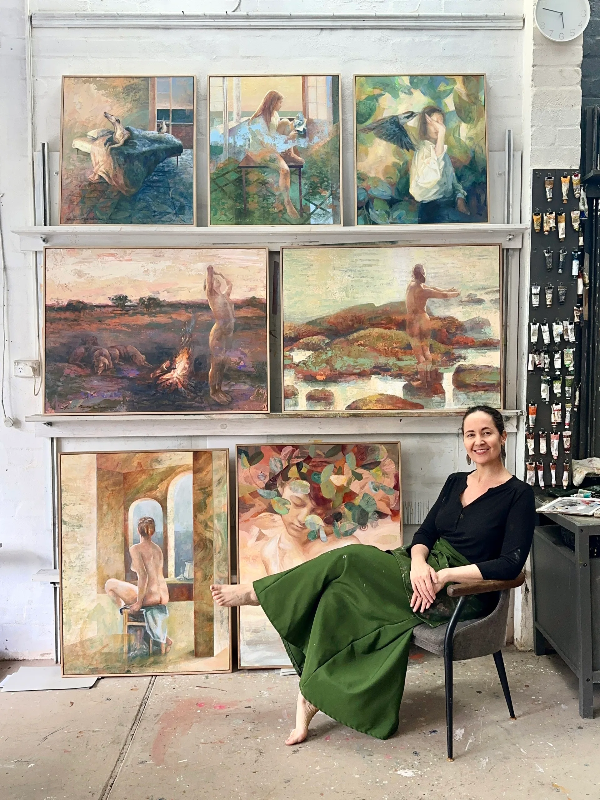Artist sitting in a studio with several colorful paintings on shelves, smiling, wearing a green skirt and black top.