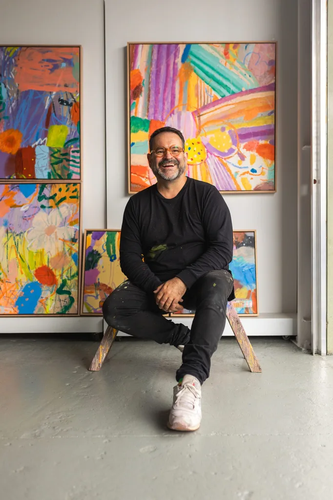 Artist in a black outfit sits smiling in front of colorful abstract paintings